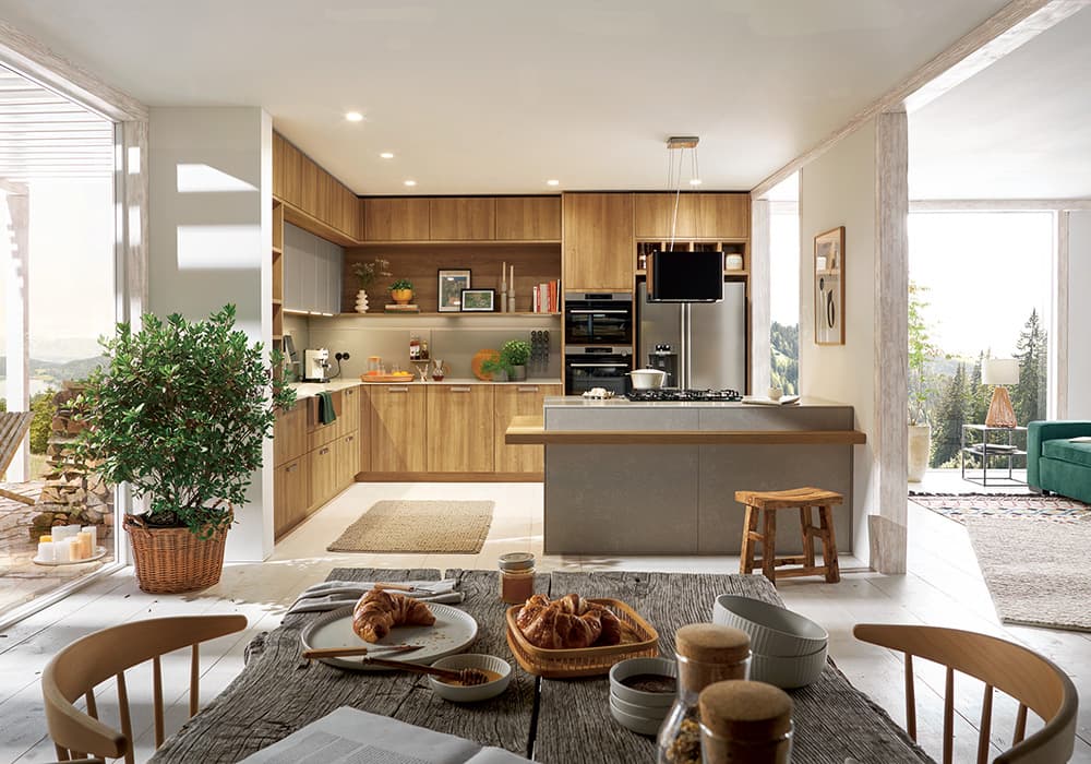 schueller Küche Bari in Holzoptik mit heller Arbeitsplatte, einem Holz-Esstisch im Vordergrund sowie hohen Fenstern mit Blick auf den Wald