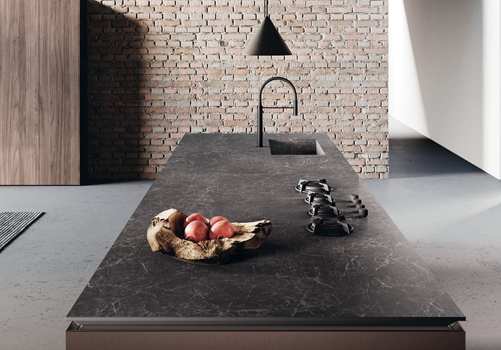 Keramik-Arbeitsplatte in dunkler Marmoroptik auf einer Kücheninsel mit integriertem Spülbecken, vier Gasplatten, einer Obst-Holzschale & einer Ziegelwand im Hintergrund