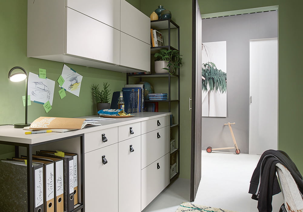 Blick aus einem Büro mit weißen Schränken & Hängeschränken sowie offenen Elementen & grüner Wandfarbe durch eine offen stehende Tür in den Flur